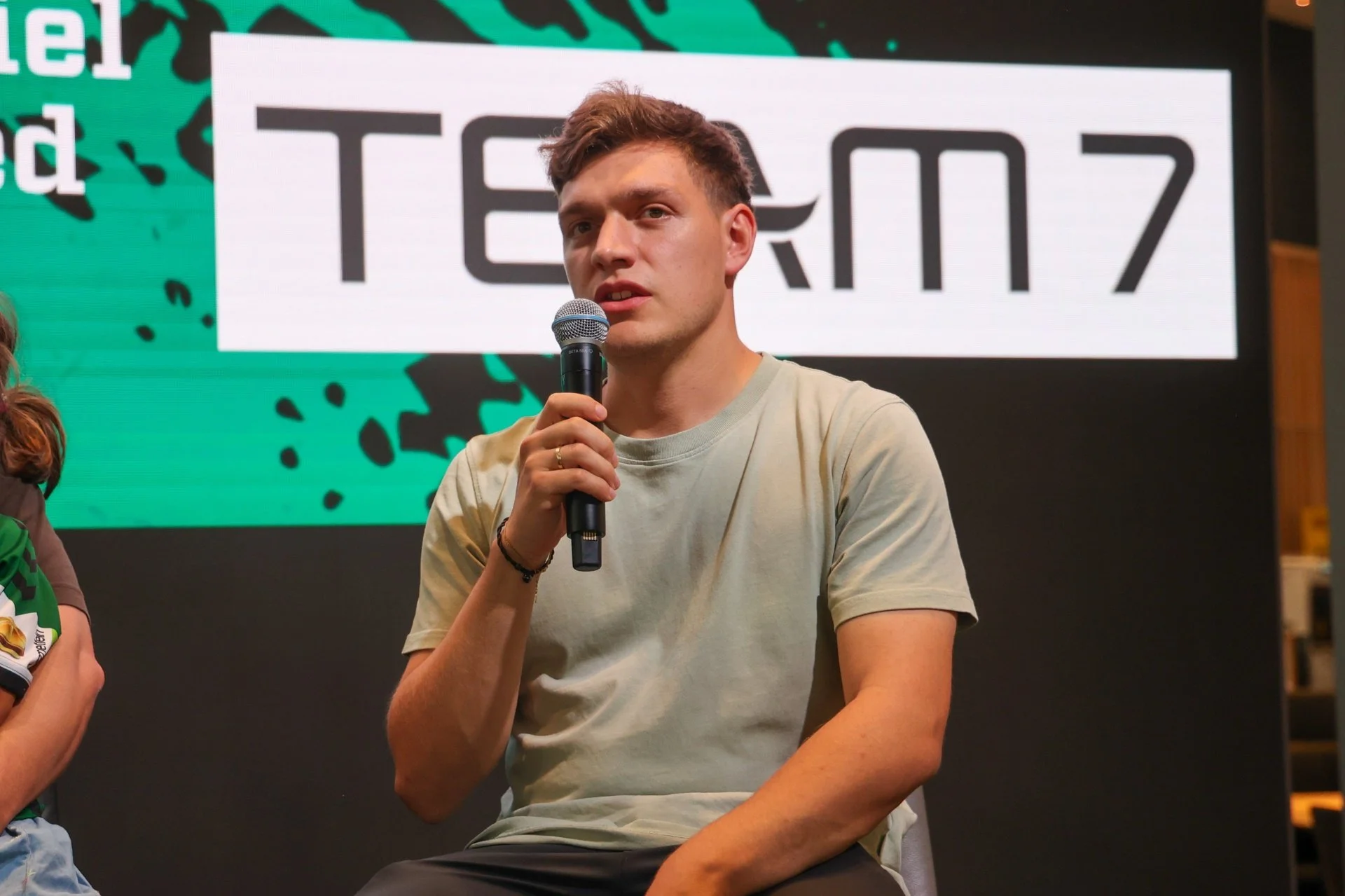 Lukas Brandl holding a microphone and speaking, sitting on a chair. He wears a light-colored t-shirt and is in front of a large digital screen displaying the word 'TEAM' in bold black letters, along with colorful abstract designs.