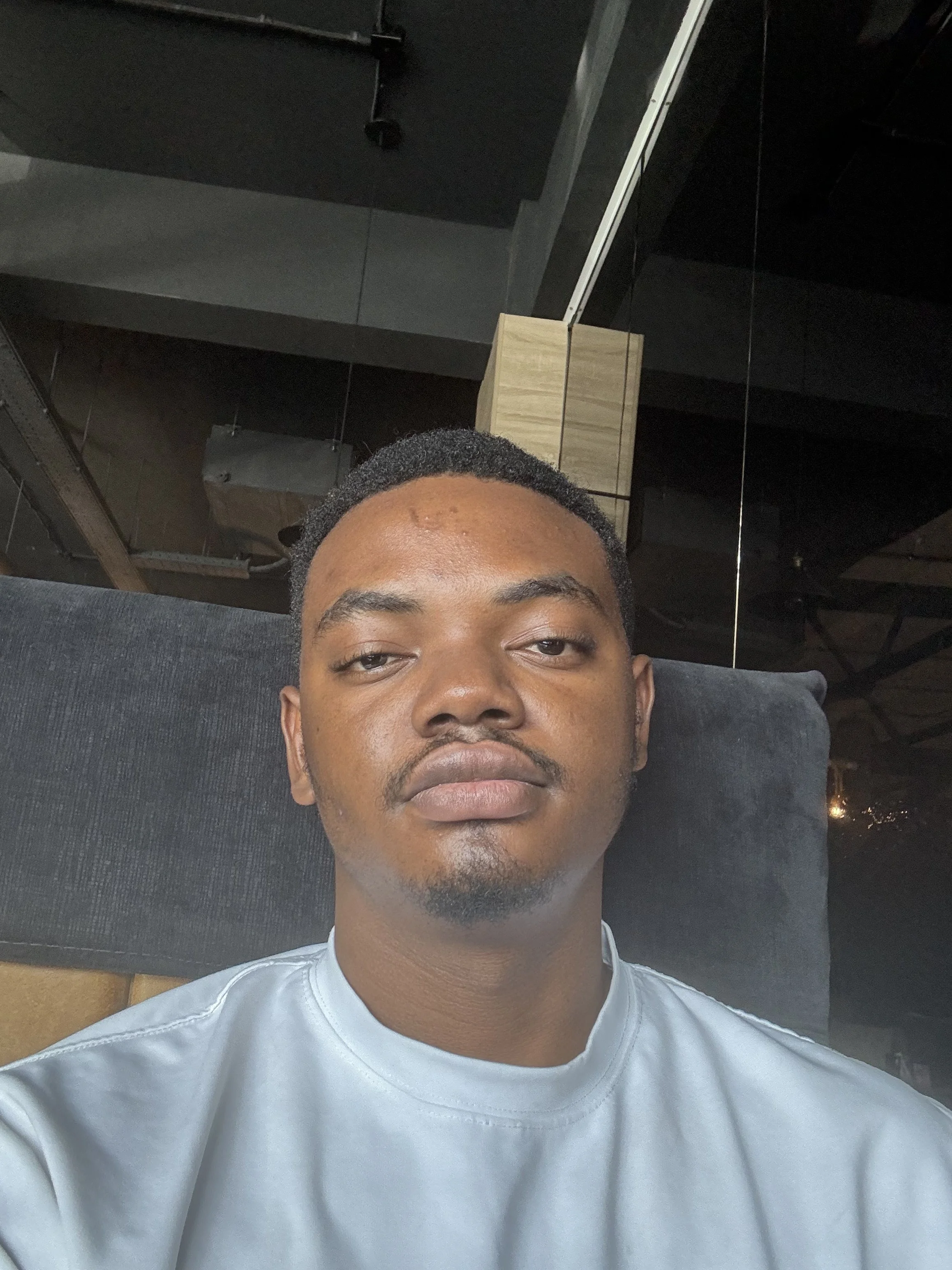 Close-up selfie of Israel .T. Agboola with short hair, looking directly at the camera, wearing a light gray shirt, with a black couch and ceiling fixtures in the background.