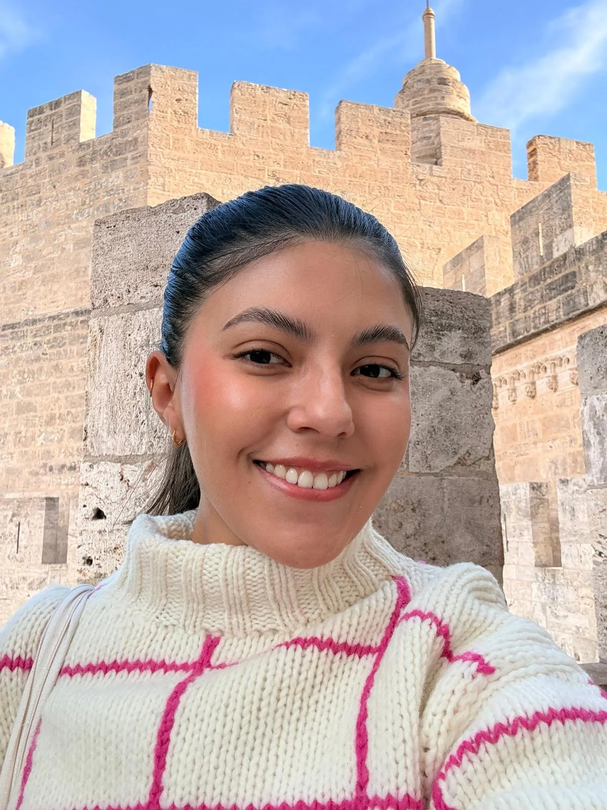 A young woman smiling in front of a historic stone castle with crenellations and a tower, under a clear blue sky.
