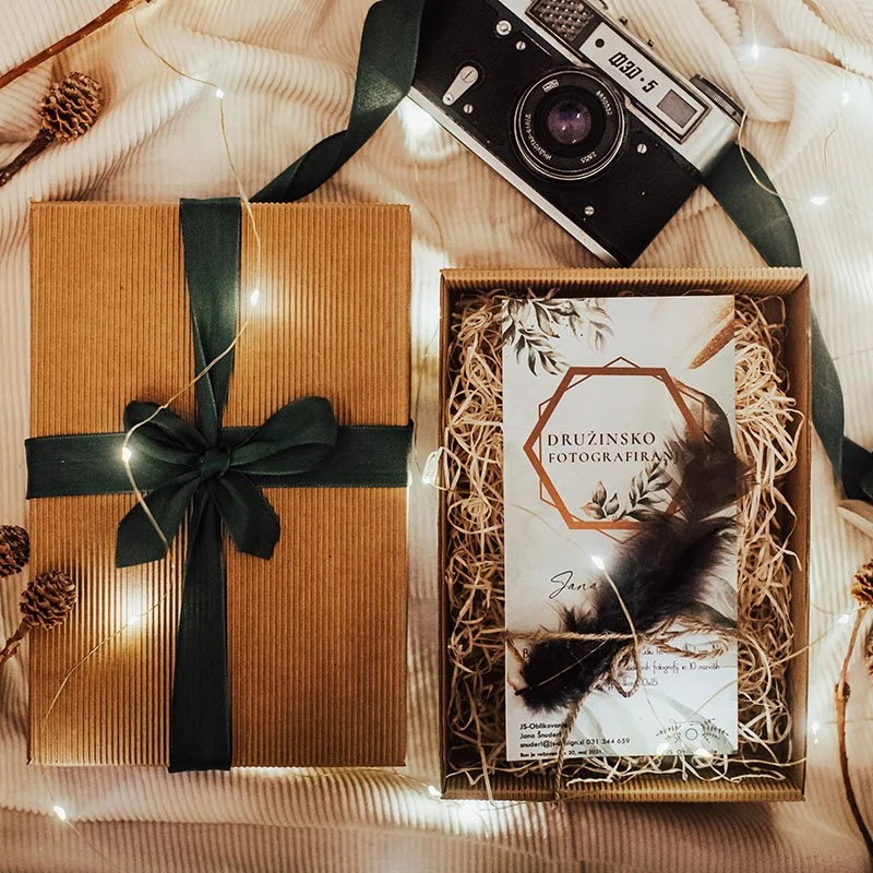 A gift box wrapped in brown paper with a dark green ribbon and bow, a cardboard box filled with shredded paper containing a business card and feather, a vintage camera, and fairy lights surrounding them on a textured fabric surface.