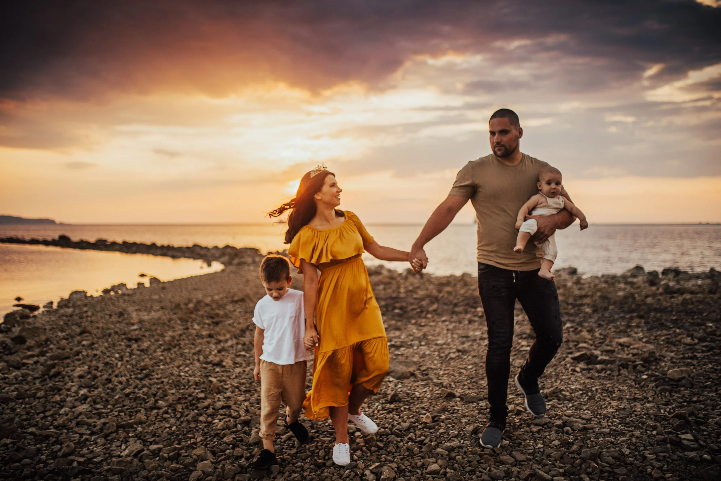 A family of four walking along a rocky beach during sunset, with the mother in a yellow dress, father carrying a baby, and two children walking beside them.