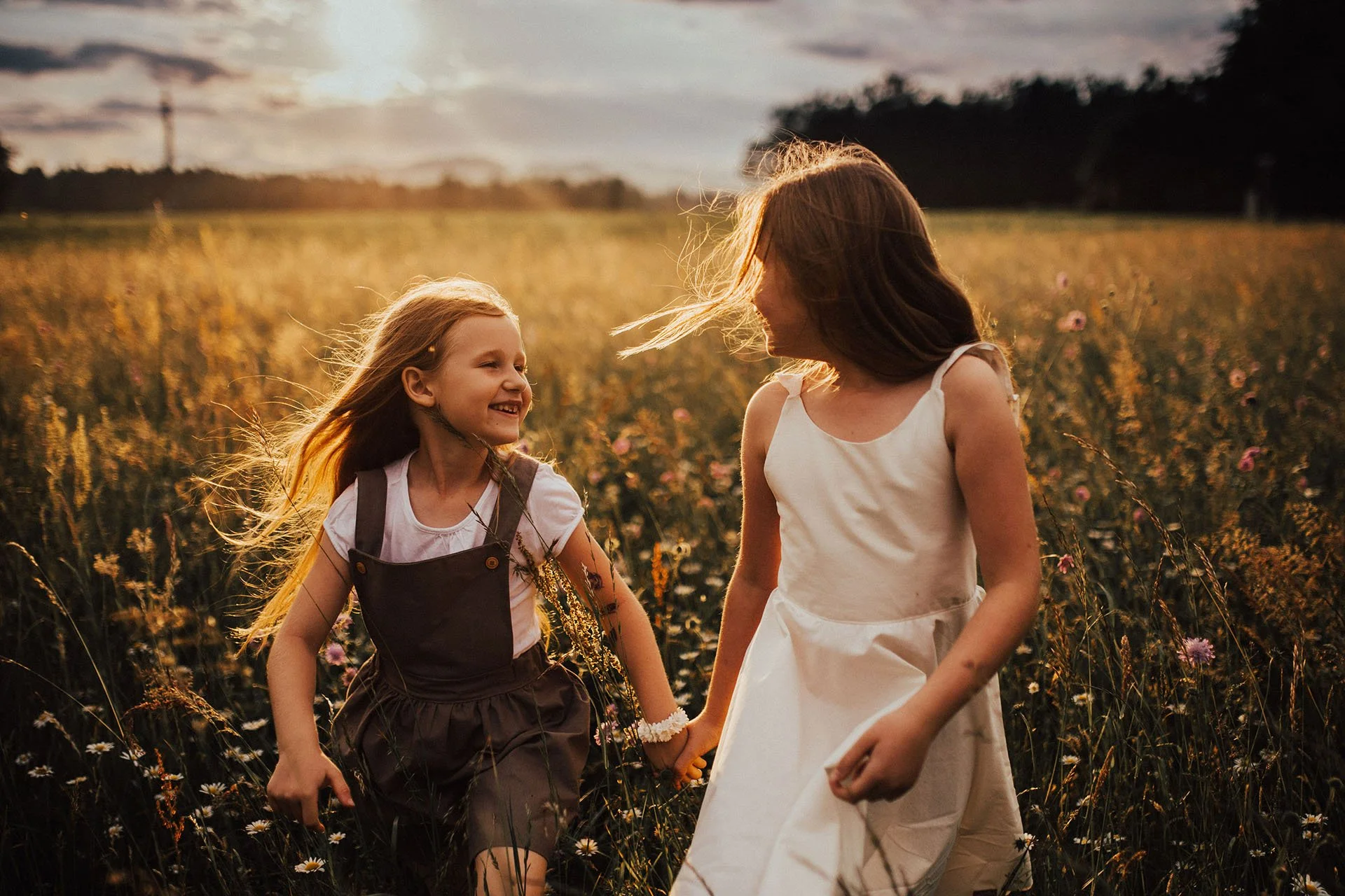 Two young girls holding hands and smiling while walking through a field of wildflowers at sunset.