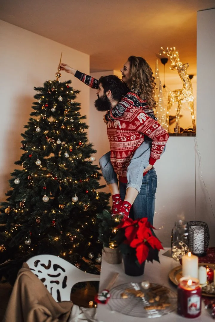 A couple decorating a Christmas tree together; the woman is on the man's back while reaching to hang an ornament.