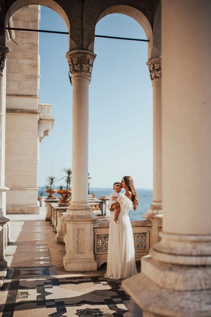 A woman in a white dress holding a young girl in a similar dress, standing on a balcony with large stone columns overlooking the sea on a sunny day.