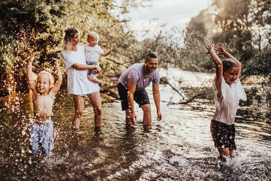 Family enjoying wading and playing in a shallow river surrounded by trees during sunset.