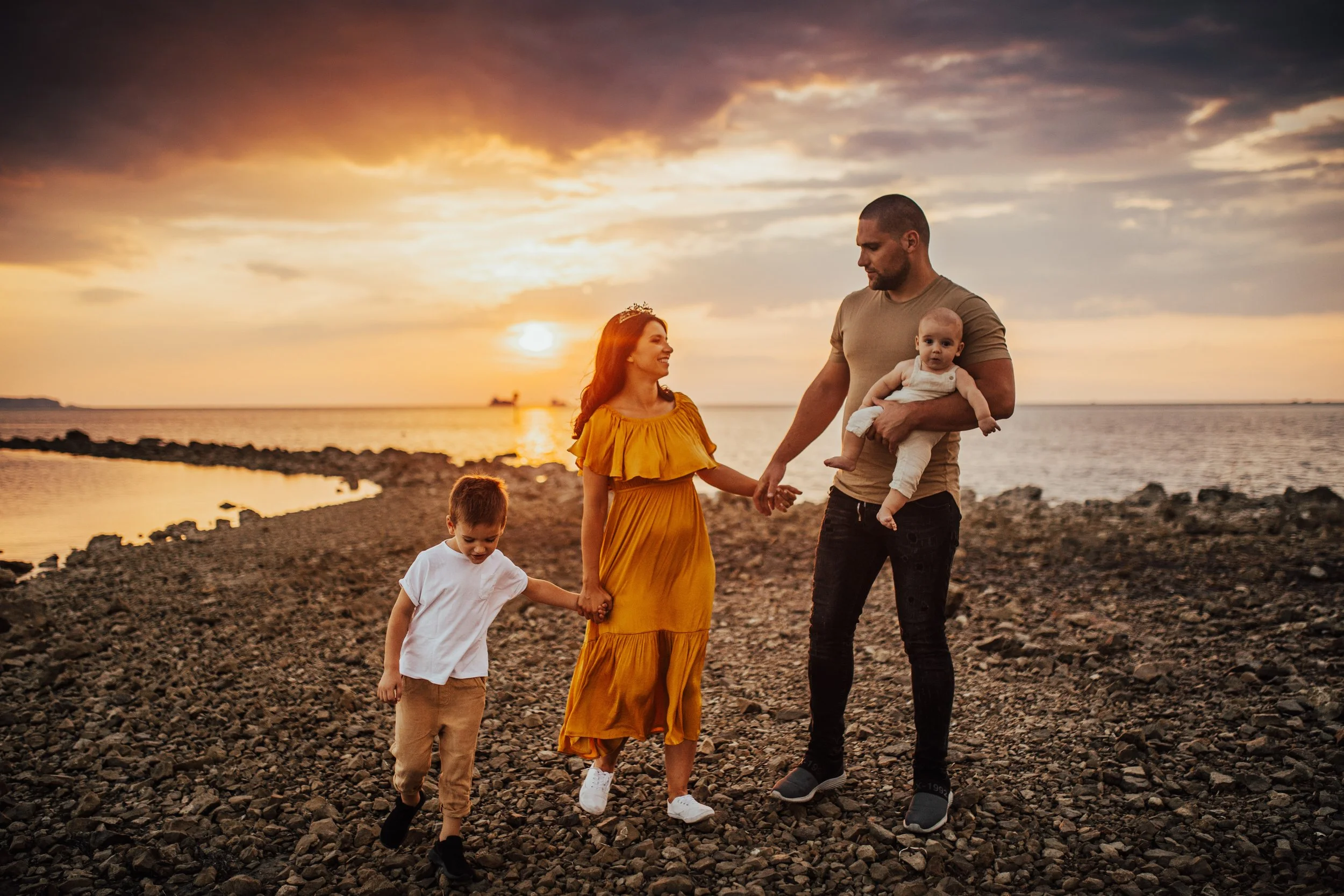 Family of four walking on a rocky beach at sunset, including a woman in a yellow dress, a man holding a baby, and a young boy with white shirt and beige pants.
