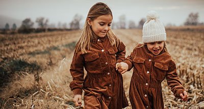 Two young girls walking together in an open field during daytime, holding hands and wearing brown overalls and knit hats.