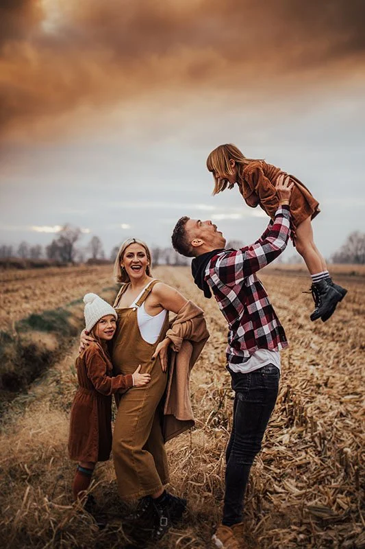 A family of four enjoying time outdoors in a field during sunset, with the father lifting a young girl in the air while the mother and another girl smile.
