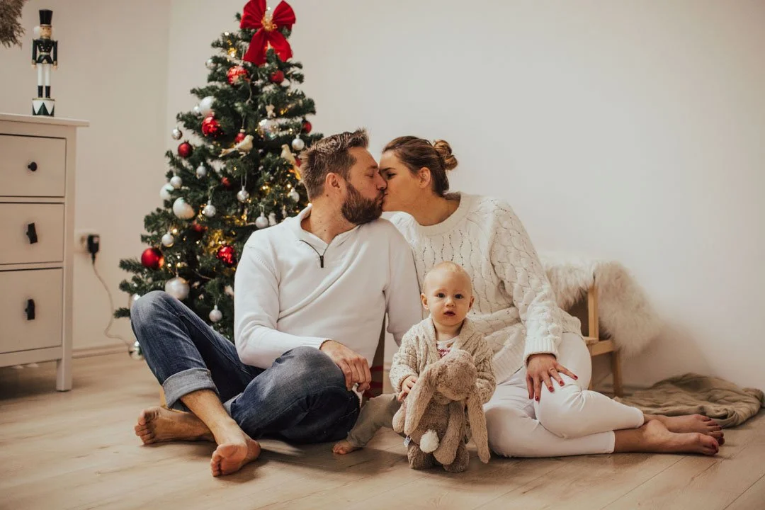 A family of three sitting on the floor in front of a decorated Christmas tree, with the parents kissing and their young child holding a stuffed toy.