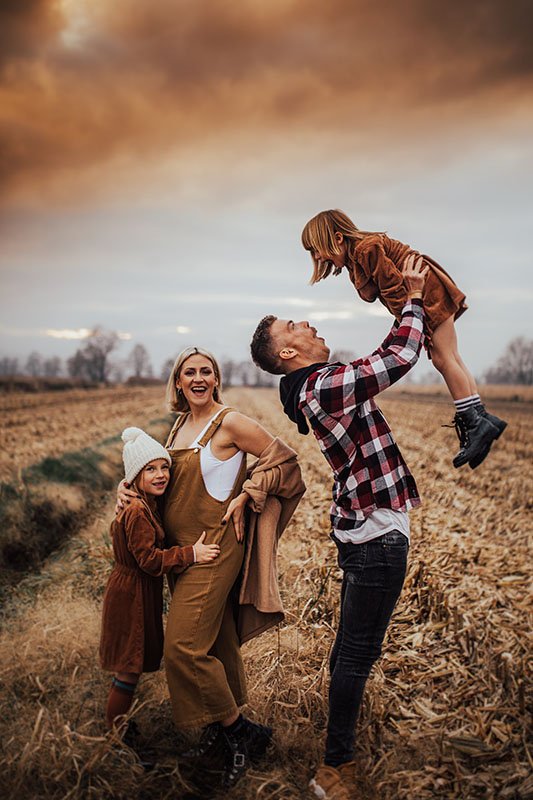 A family of four outdoors in a field during sunset, smiling and playing together. The father lifts a young girl, while the mother and another child smile nearby.