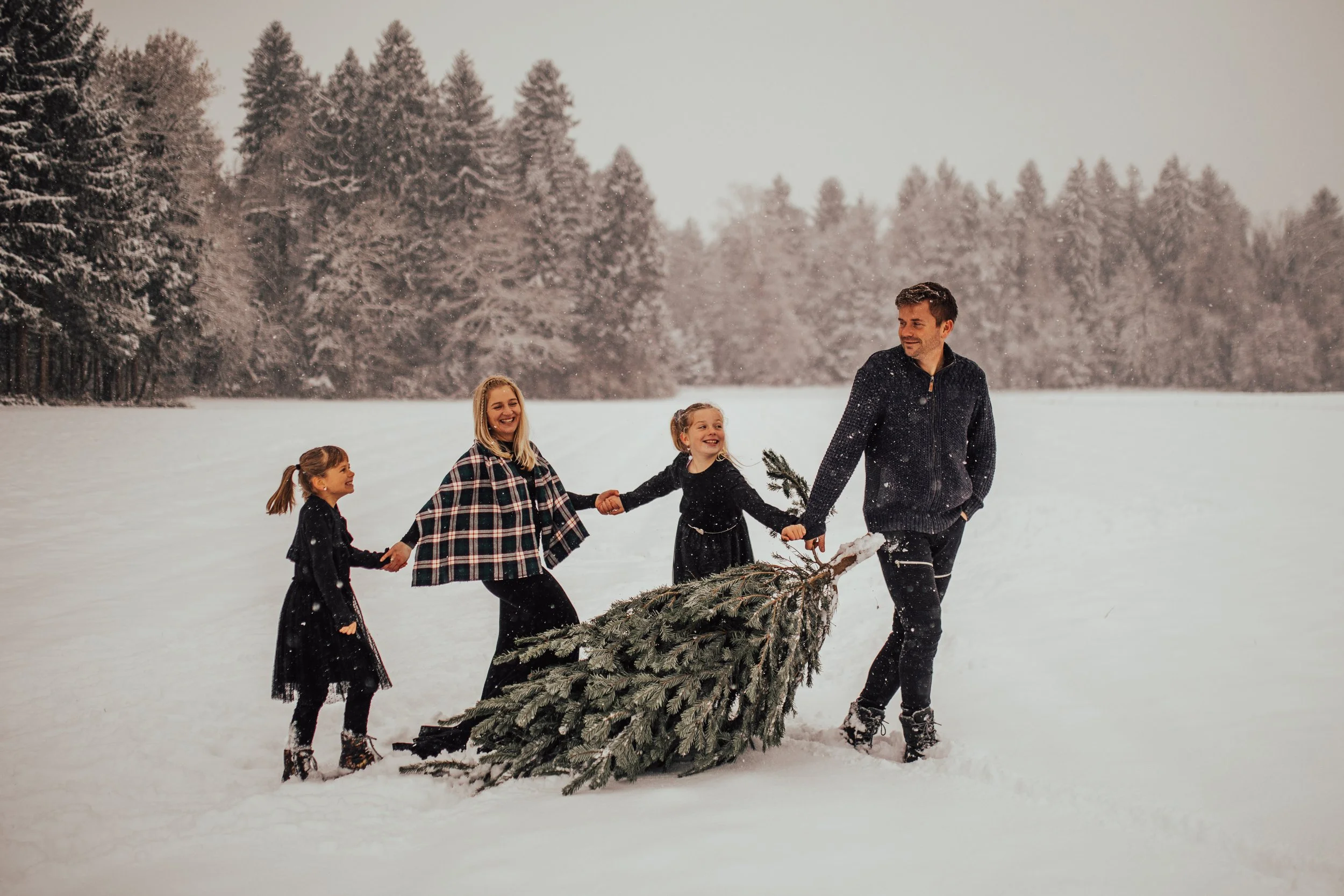 A man and four girls are outside in a snowy field, holding a pine tree and its branches. They are dressed warmly and smiling, surrounded by a forest of snow-covered trees.