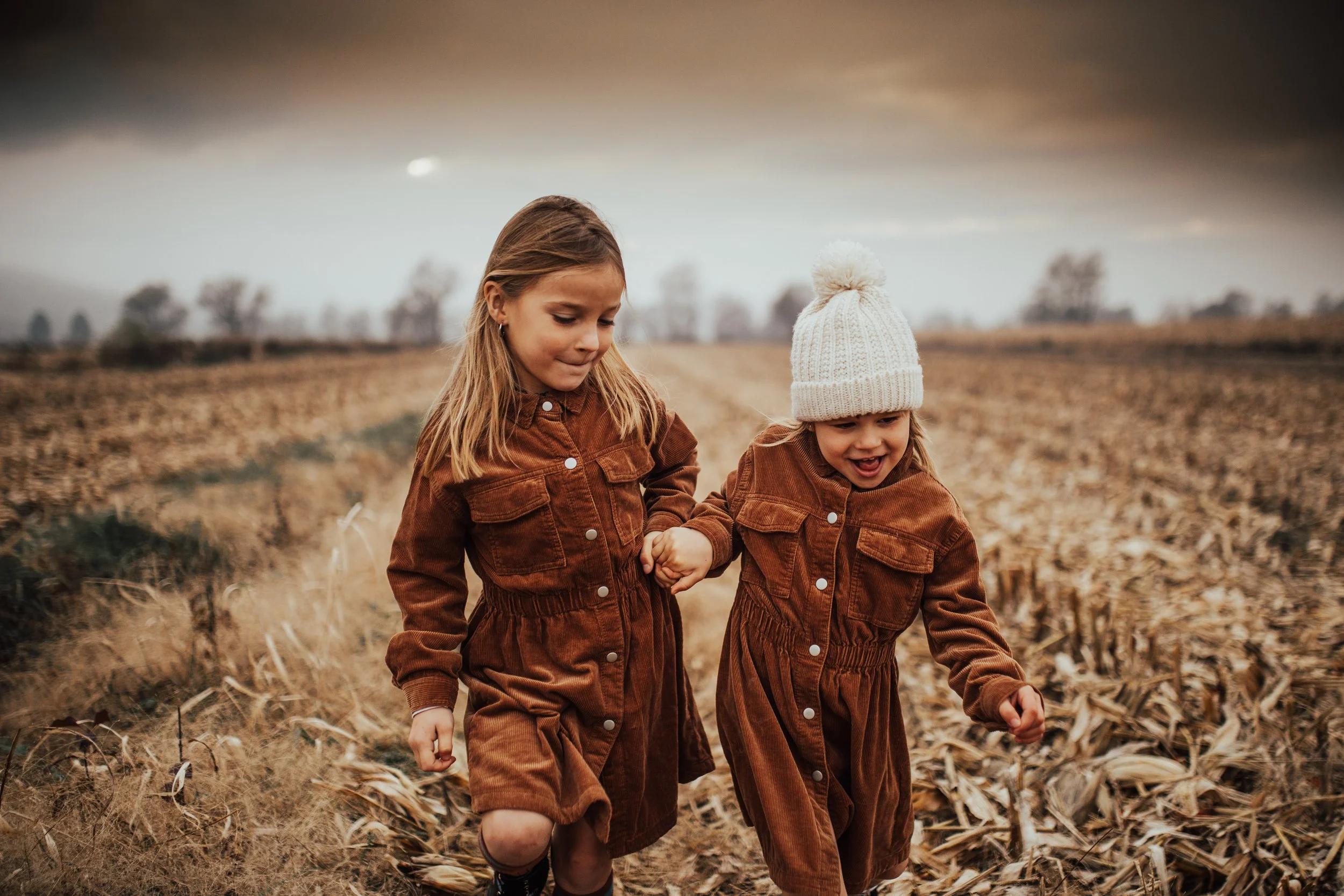 Two young girls, one with long blonde hair and the other with long hair, wearing brown jackets and walking hand in hand in a harvested cornfield on a cloudy day.