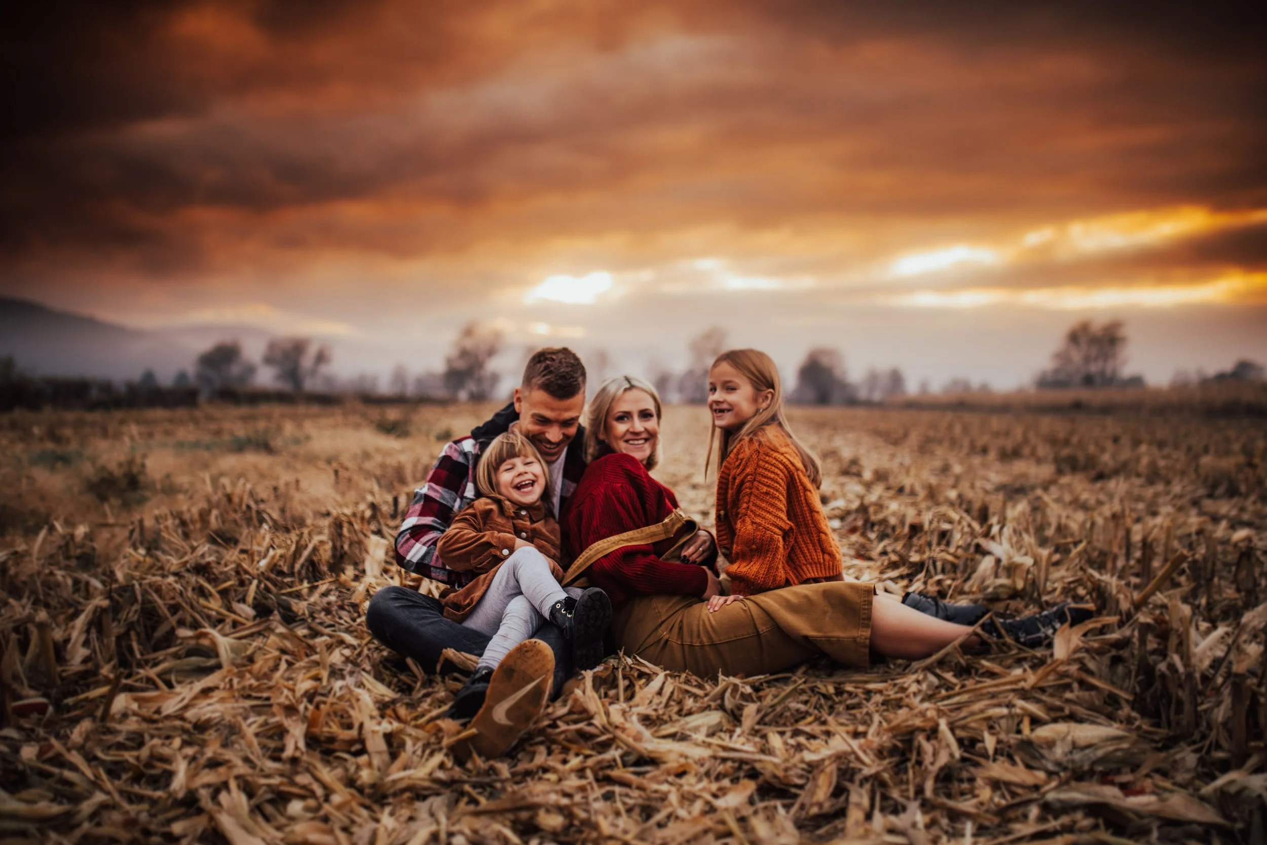 A family of four, including two children, sitting on a fallen leaf-covered ground in a harvested field during sunset, smiling and enjoying each other's company.