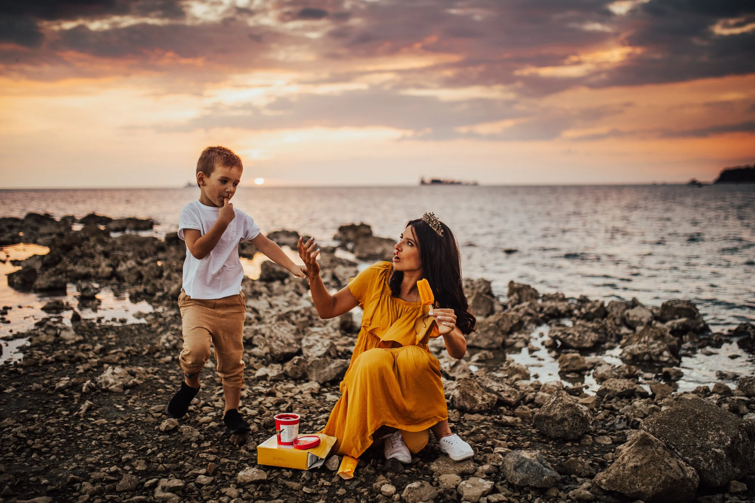 A woman and a young boy on a rocky beach at sunset. The woman is kneeling and wearing a yellow dress, holding a small object, and the boy is standing, touching his mouth. The sky is colorful with orange, pink, and purple clouds over the ocean, with a