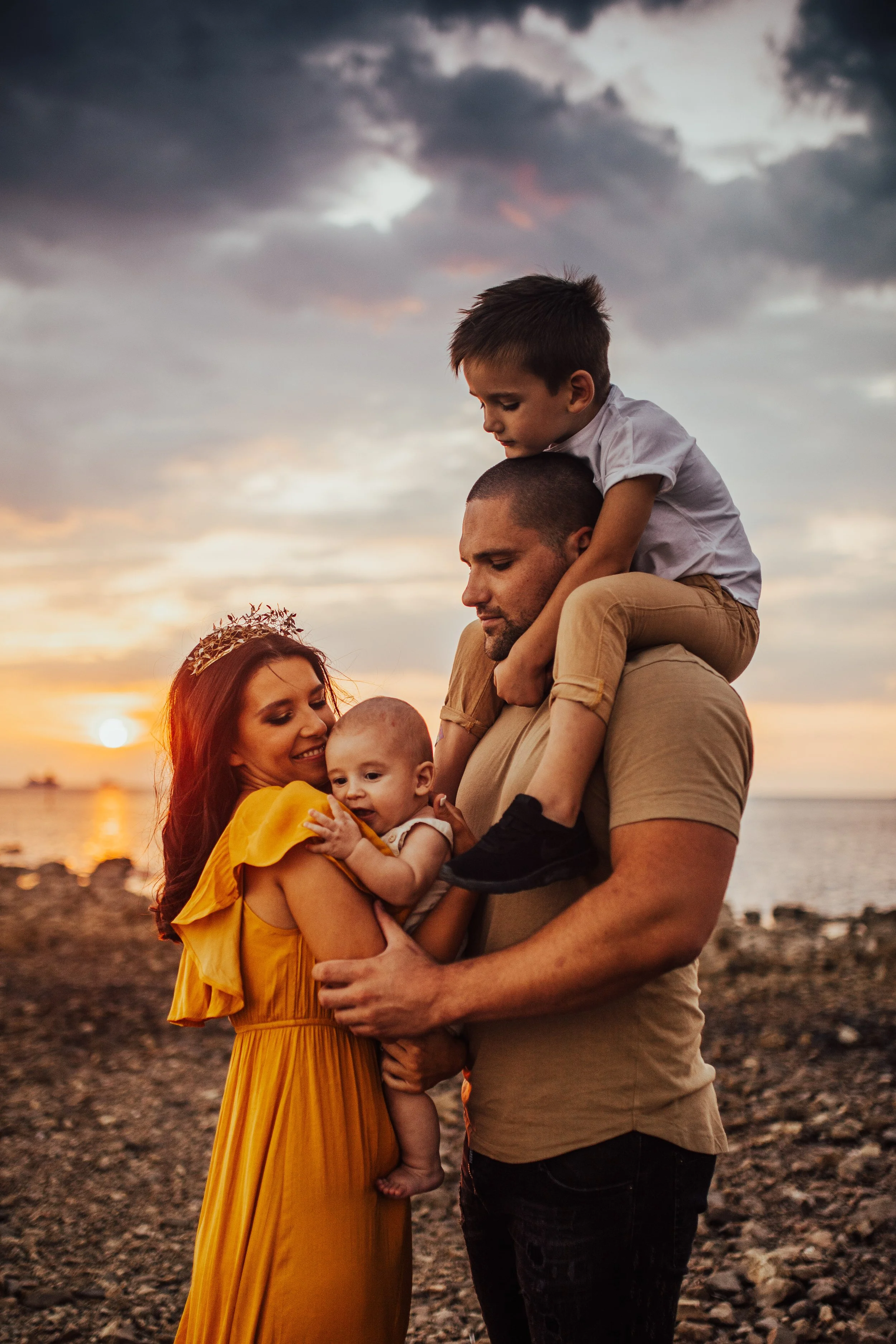 A family of four enjoying a sunset at the beach. The mother, with red hair and a tiara, holds a baby. The father, with a shaved head, carries a young boy on his shoulders. The sky is partly cloudy with orange and purple hues, and the ocean is visible in the background.