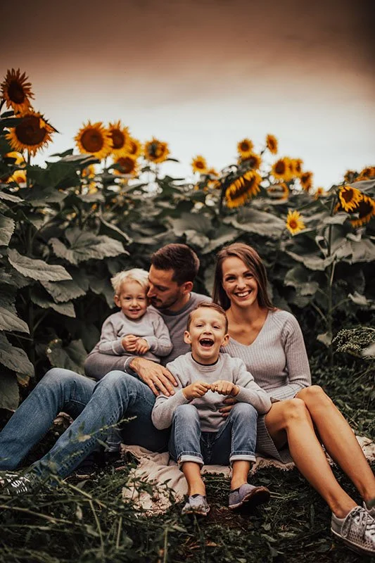 A family of four sitting on the ground in a sunflower field, smiling and enjoying each other's company during sunset.