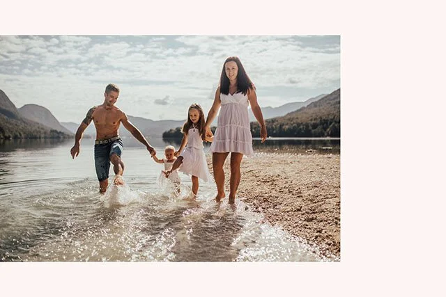 A family of four walking along a lake's shoreline with mountains in the background, holding hands and splashing water, during daytime.