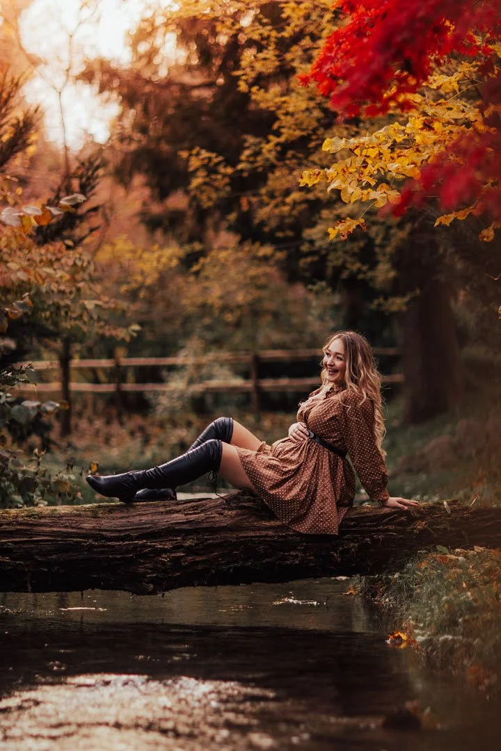 A woman with long wavy hair sitting on a fallen log over a stream in an autumn forest, smiling and looking to the right, with red, orange, and yellow leaves overhead.