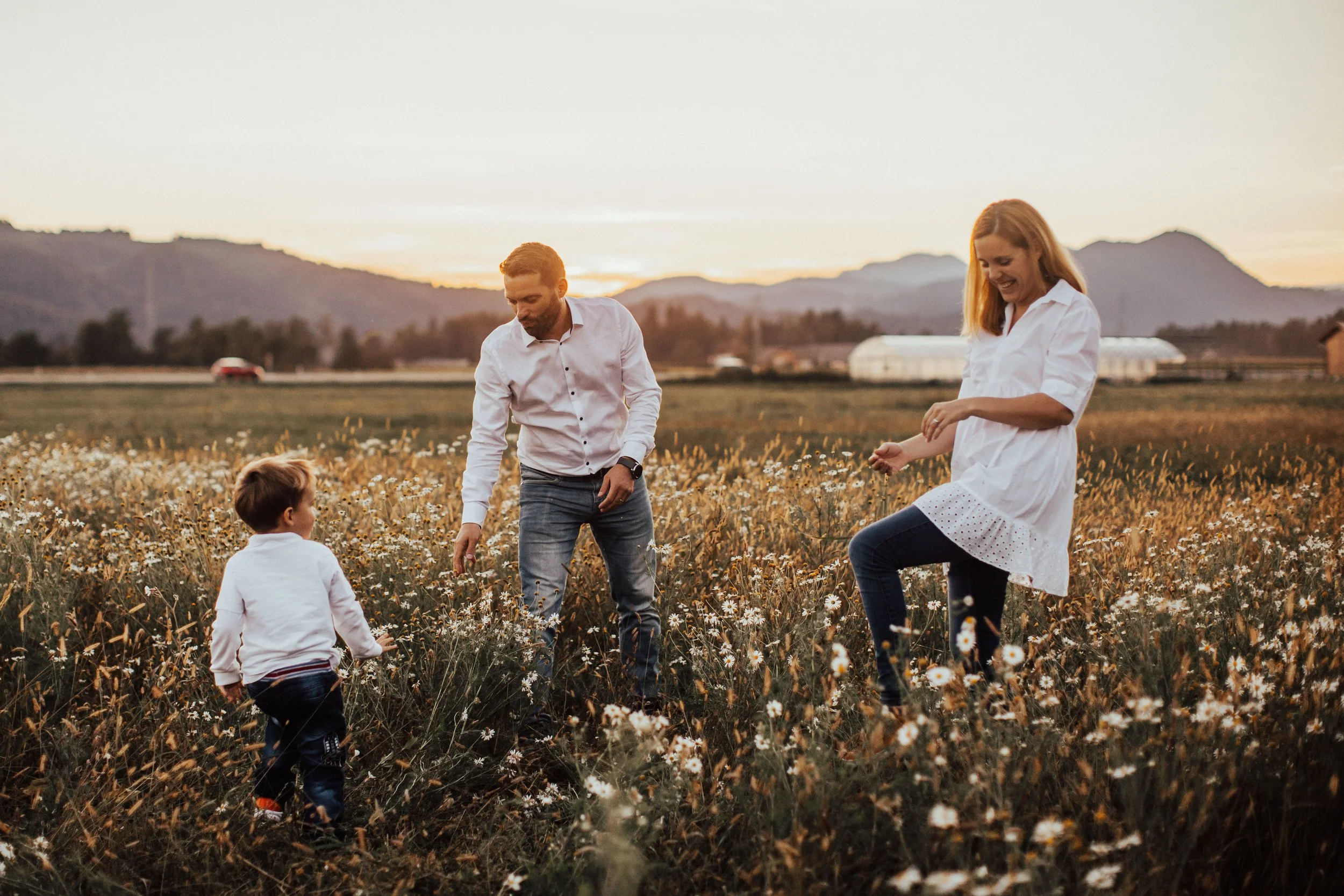 A family of three in a field of wildflowers during sunset. The father and mother are standing while the young son is kneeling. They are all dressed casually and smiling as they enjoy the outdoor activity.