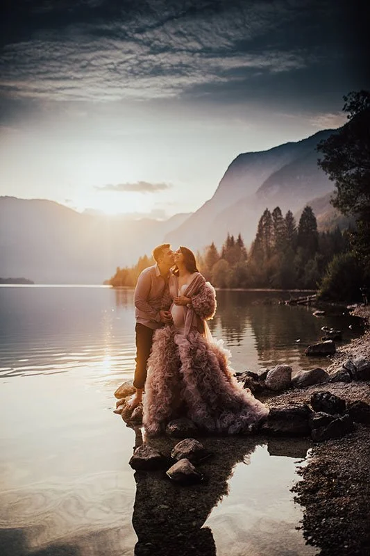 A couple dressed in formal attire kissing on rocks by a lake at sunset, with mountains and trees in the background.