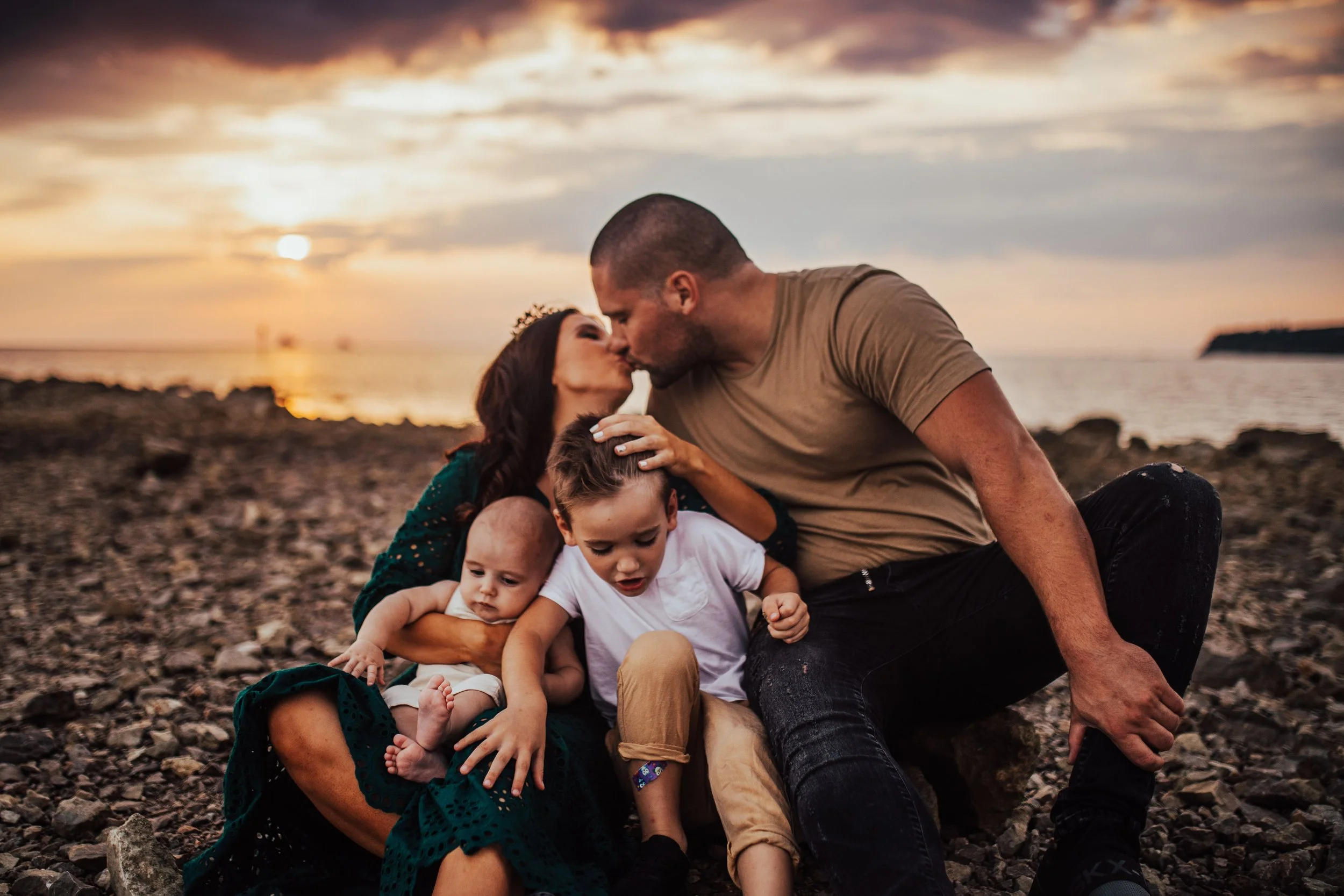 A family of four sitting on a rocky beach during sunset, sharing a kiss and hugs.