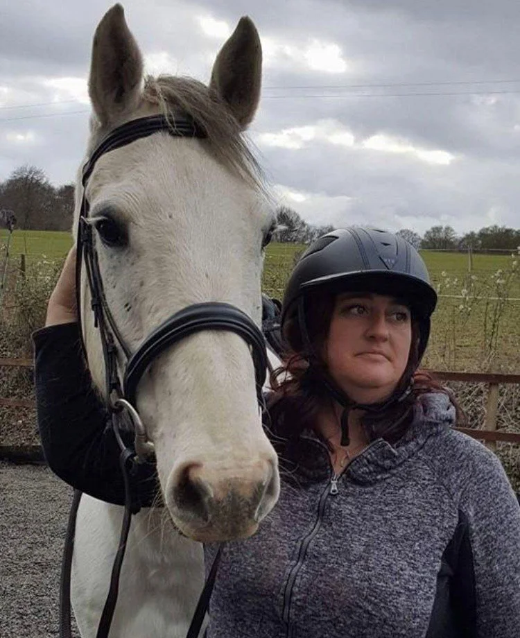 A woman wearing a riding helmet standing next to a white horse with black bridle in an outdoor rural setting overcast with clouds.
