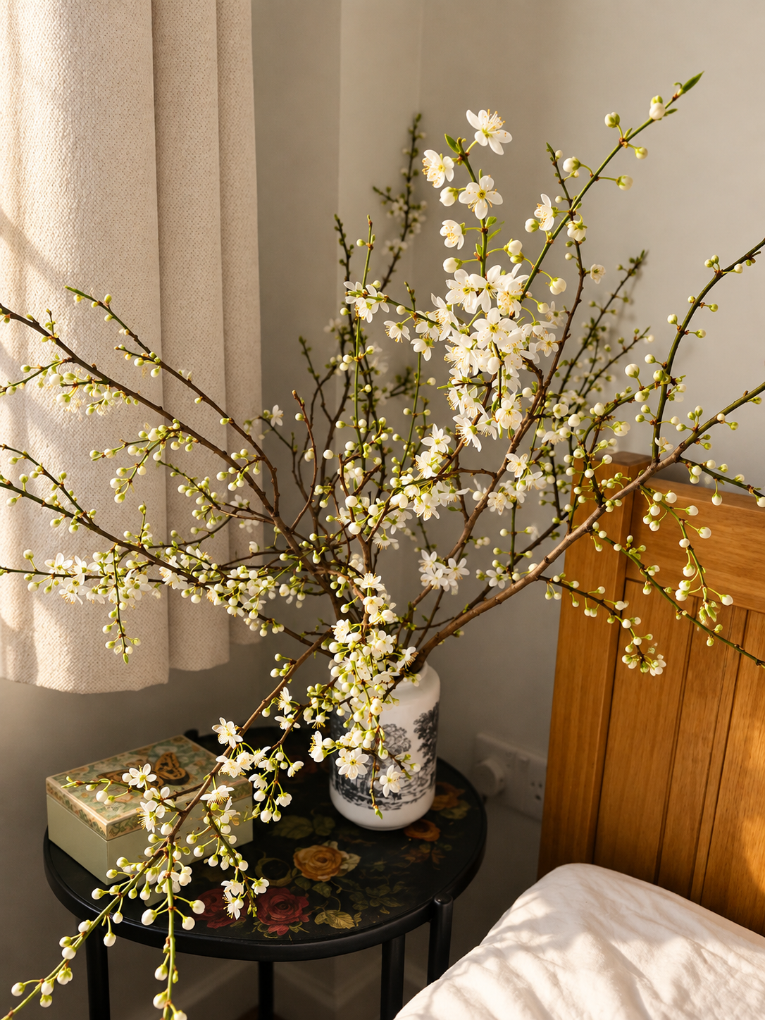 A large branch of white flowering blossoms in a white vase on a round black side table next to a bed with white bedding and beige curtains in a cozy bedroom.