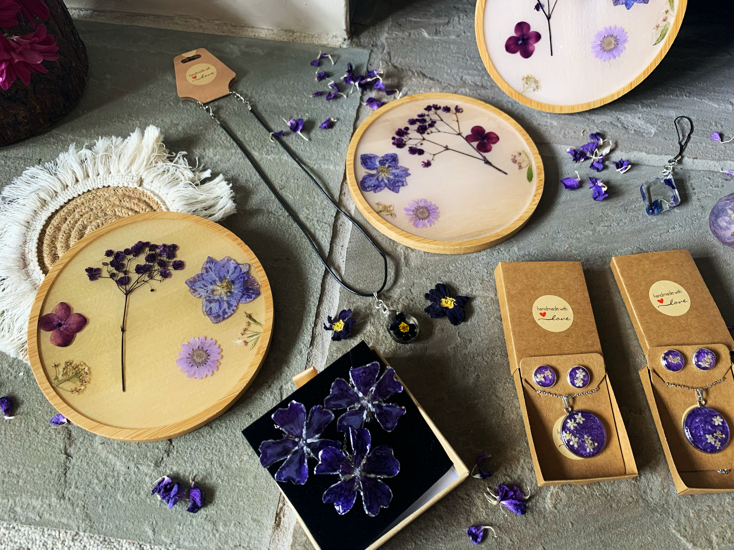 Display of jewellery and decorative items including real dried purple flowers on wooden coaters,  boxes with purple flower jewellery, and jewellery pieces with purple flowers on small paper packaging at a market stand.