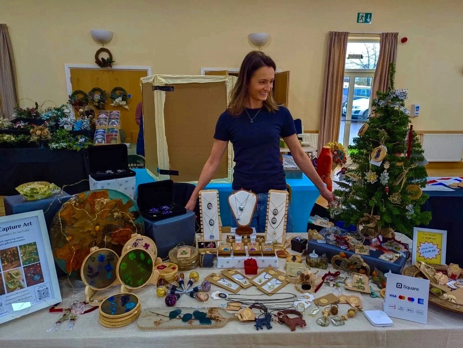 A woman stands behind a table filled with jewelry and craft items at a craft fair, with Christmas decorations and a Christmas tree in the background.