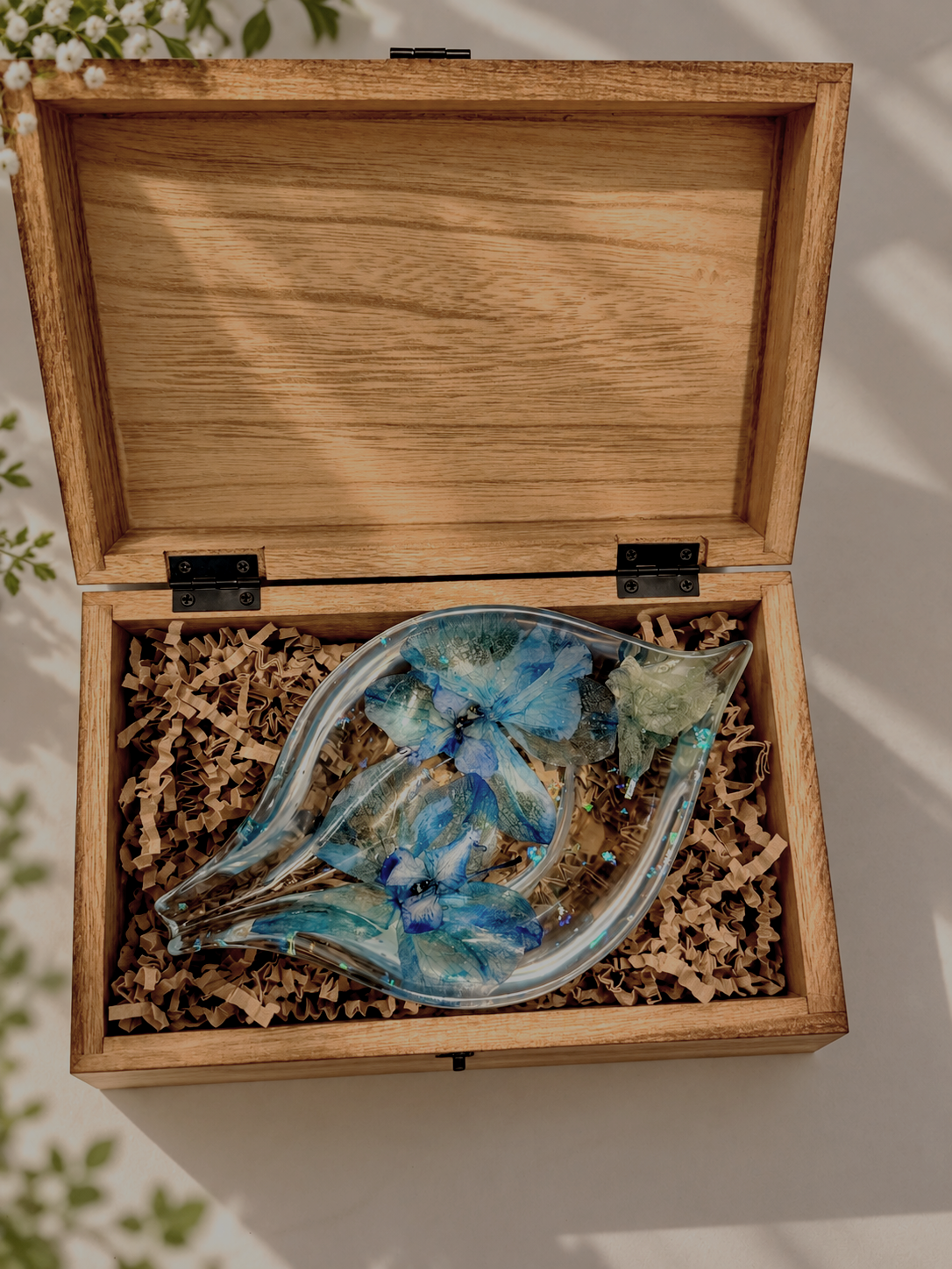 A wooden box containing a glass sculpture decorated with blue butterfly and floral designs, set on shredded paper.