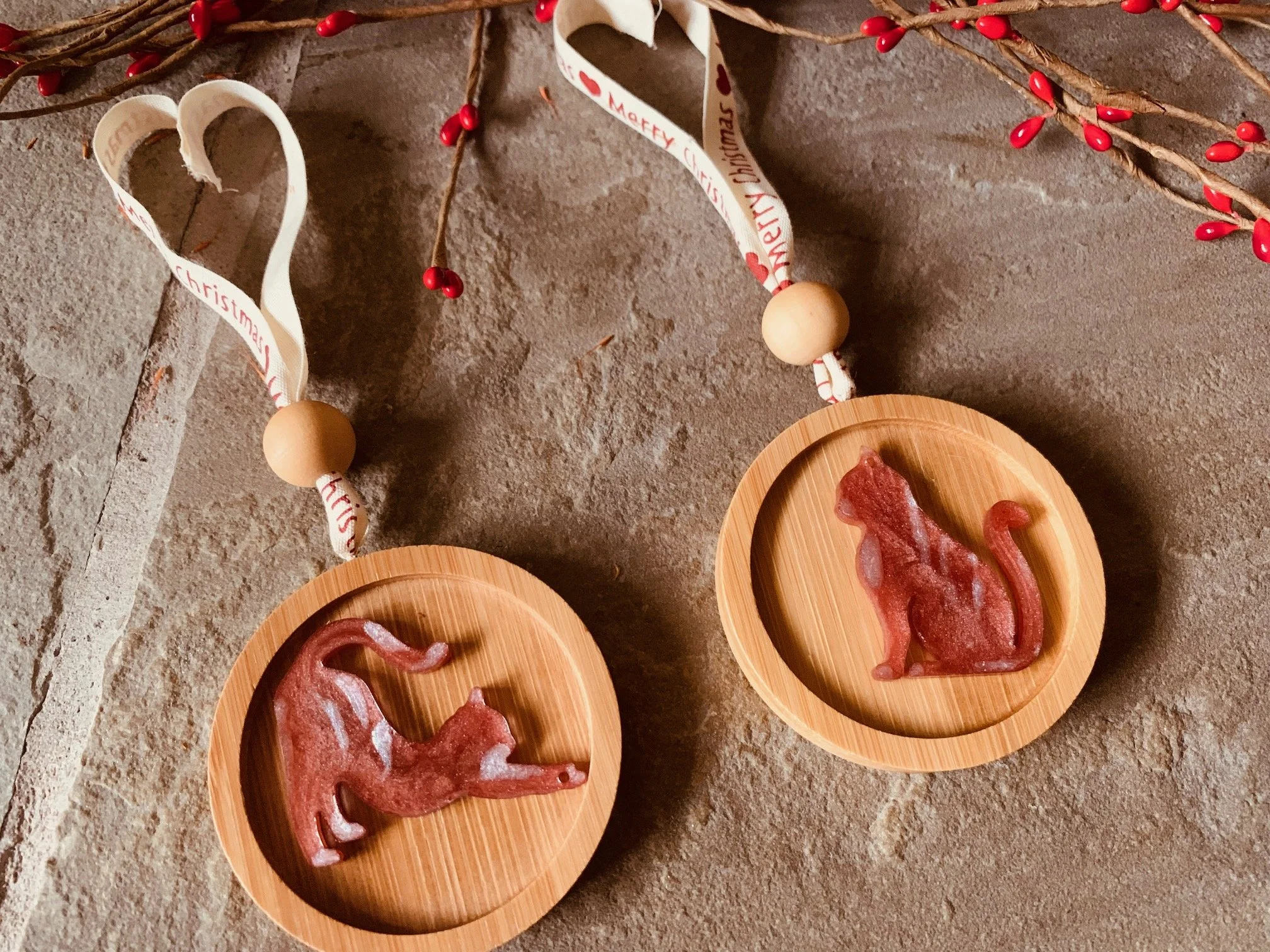 Two handmade ginger red resin cat Christmas decorations on round wooden ornaments, featuring sitting and stretching cat designs, festive ribbons and red berry branches.