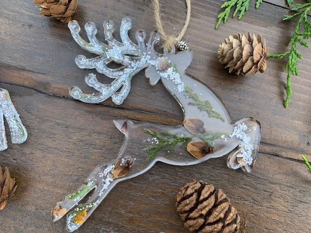 Handmade clear resin Christmas reindeer with intricate antlers, real preserved greenery, natural pine cone pieces and silver snow like sparkle on rustic wood.