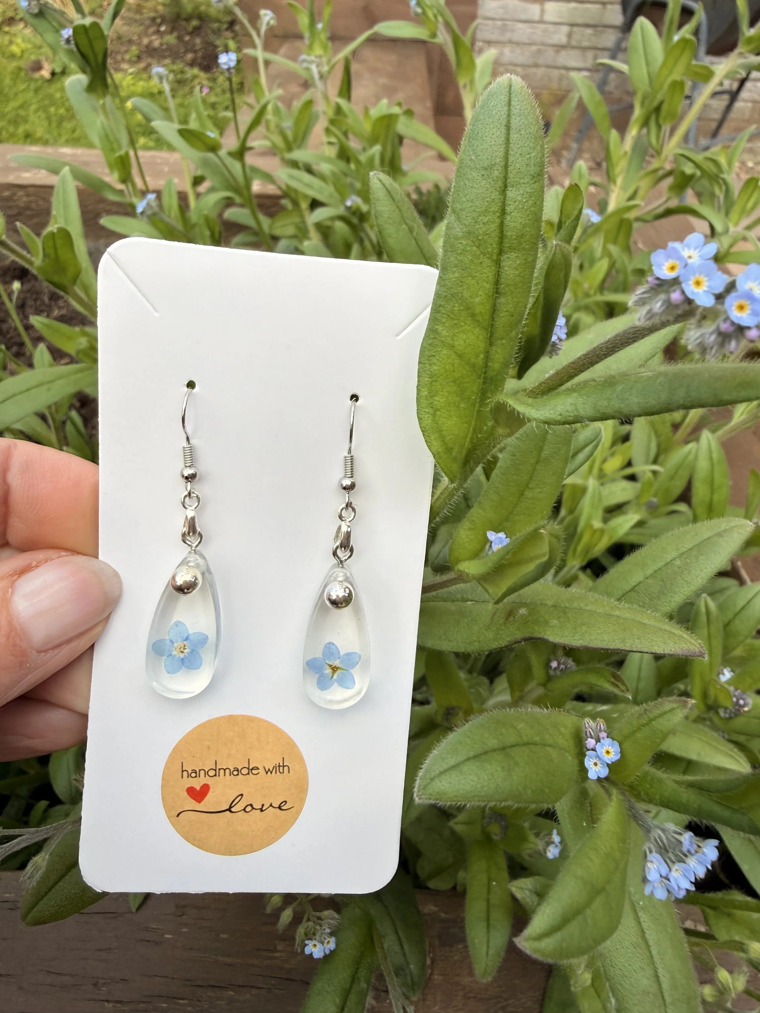 A pair of silver and clear glass teardrop earrings with small blue flowers inside, displayed on a white earring card with a sticker that reads "handmade with love." The background consists of green leaves and small blue flowers of a plant.