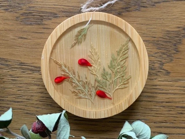 Close up a handmade wooden Christmas ornament featuring real preserved greenery and decorative red berries beneath clear resin, with a natural rustic finish.