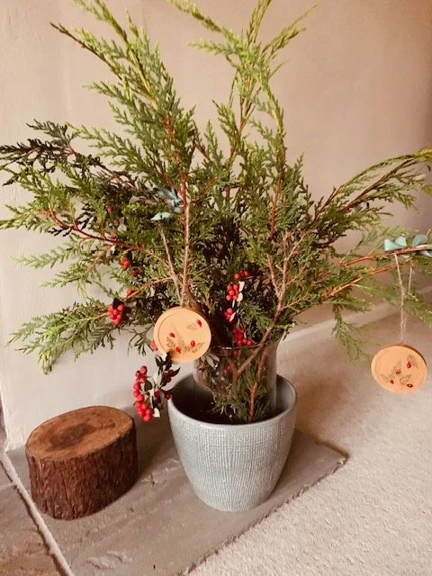 Rustic wooden Christmas tree ornament hanging among green branches, decorated with real preserved foliage, artificial red berries and a natural twine cord.