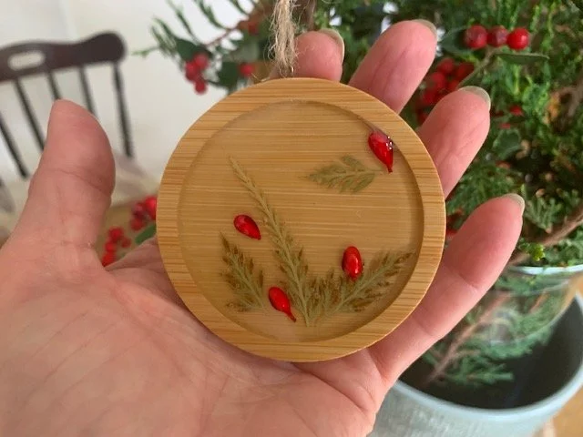 Handmade 6.5cm round wooden Christmas decoration held ina hand to show its size, featuring real preserved greenery and decorative red berries beneath clear resin.