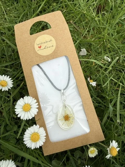 Handmade real daisy flower resin necklace in kraft gift packaging, displayed on grass surrounded by fresh daisies.