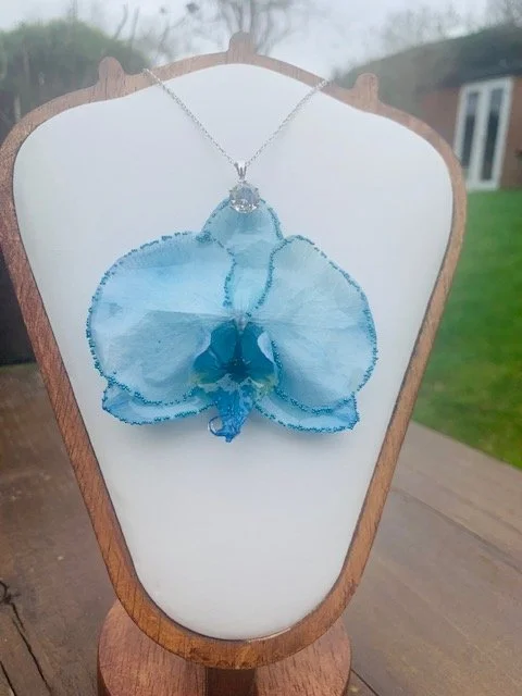 Front view of handmade real blue orchid necklace with delicate petals coated in clear UV resin, displayed on a white jewellery stand.