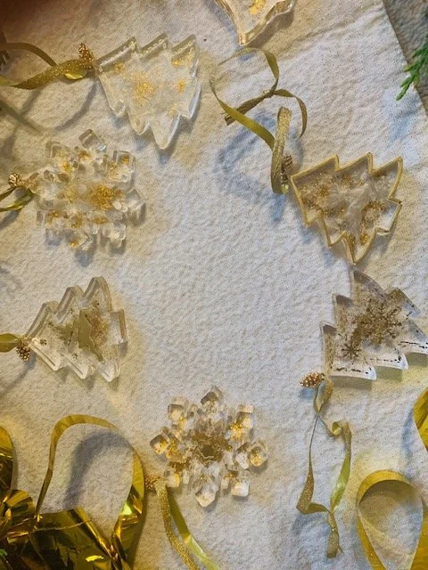 Handmade clear resin Christmas decorations featuring gold glitter details, Christamas trees, bell and snowflake designs with gold hanging ribbons.