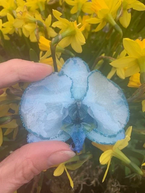 Close up of a handmade blue orchid flower necklace preserved with clear UV resin, held against yellow garden flowers.