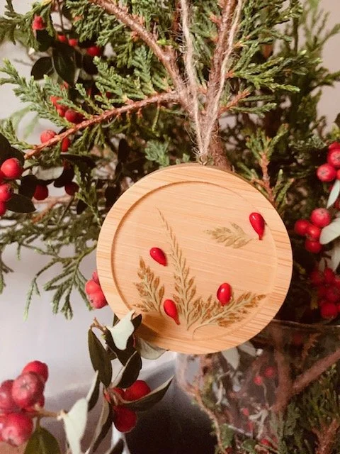 Handmade wooden botanical Christmas ornament hanging among evergreen branches, featuring real preserved greenery, decorative red berries and natural twine.