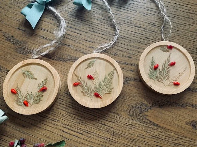 Collection of handmade round wooden Christmas tree ornaments featuring real preserved greenery, decorative red berries and rustic natural hanging cords.