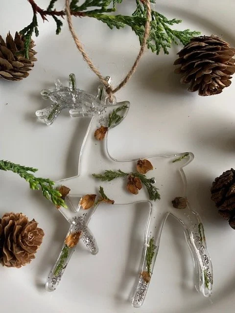 Handmade clear resin reindeer Christmas ornament featuring real greenery, nature pine cone pieces and silver winter accents, photographed in a rustic woodland setting.
