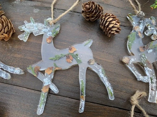 Handmade botanical resin reindeer Christmas decoration with real preserved greenery, natural pine cone pieces and shimmering silver details, displayed on rustic wood.