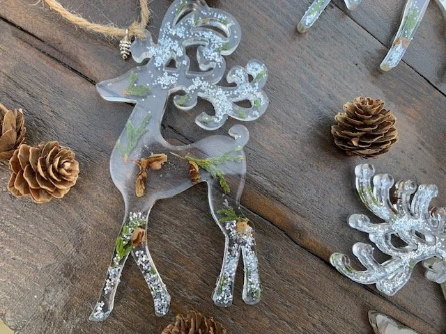 Handmade clear resin reindeer Christmas decoration with real preserved greenery, natural pine cone pieces and silver winter sparkle, displayed on rustic wood