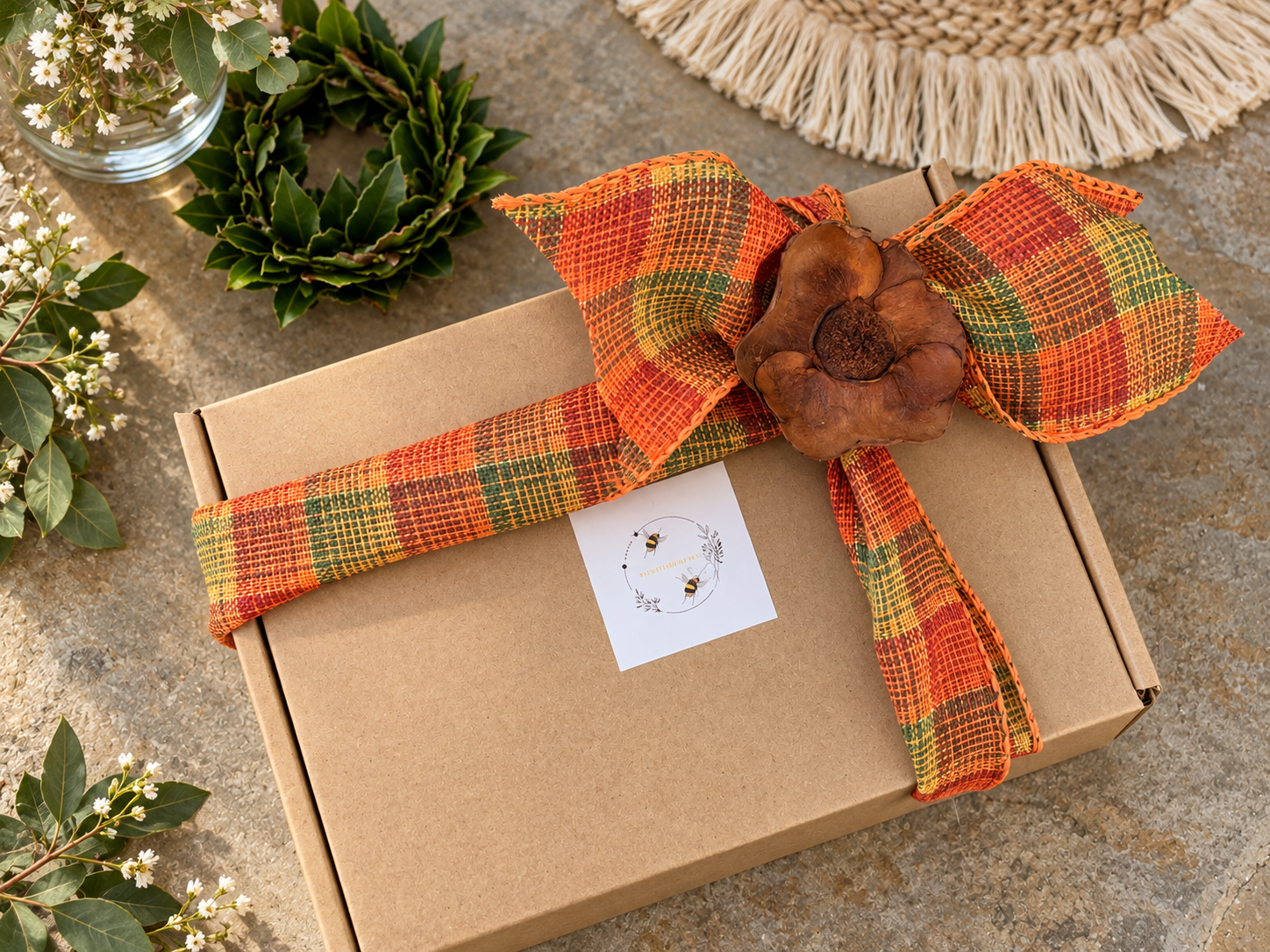 A gift box wrapped with a plaid orange, green, and yellow ribbon, adorned with a large brown flower, placed on a stone surface with green leafy plants nearby.