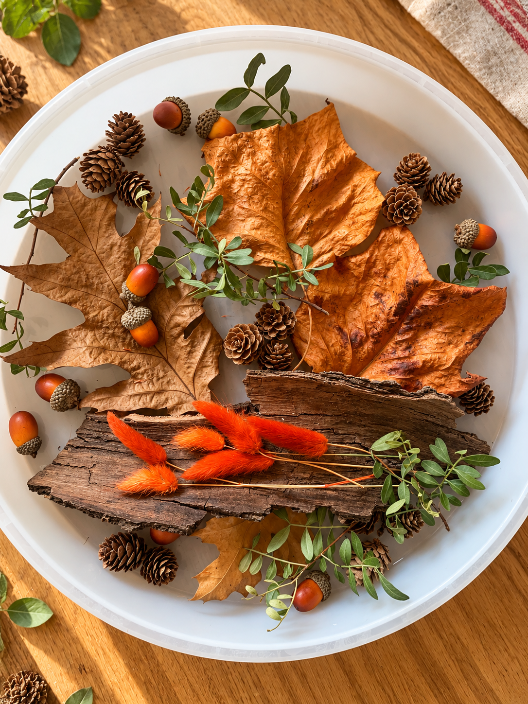 A white circular tray with fall-themed decorations including large dried orange leaves, small pinecones, orange acorns, green leaves, orange fluffy dried flowers, and a piece of bark.