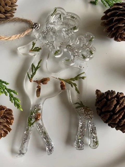 Handmade botanical resin reindeer Christmas ornament featuring real dried greenery, natural pine cone pieces and shimmering silver accents, photographed with woodland decorations.