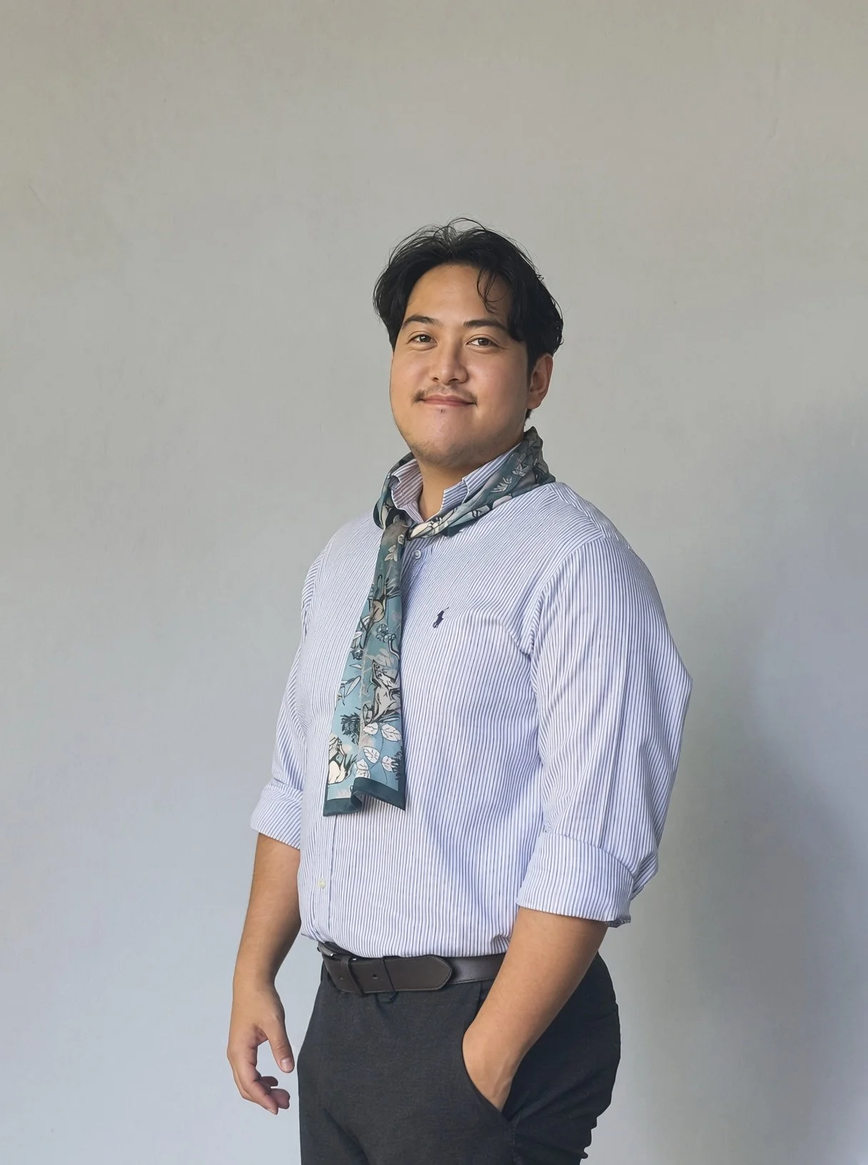 A young man with dark hair, wearing a striped dress shirt and a floral scarf around his neck, standing against a plain light-colored background.