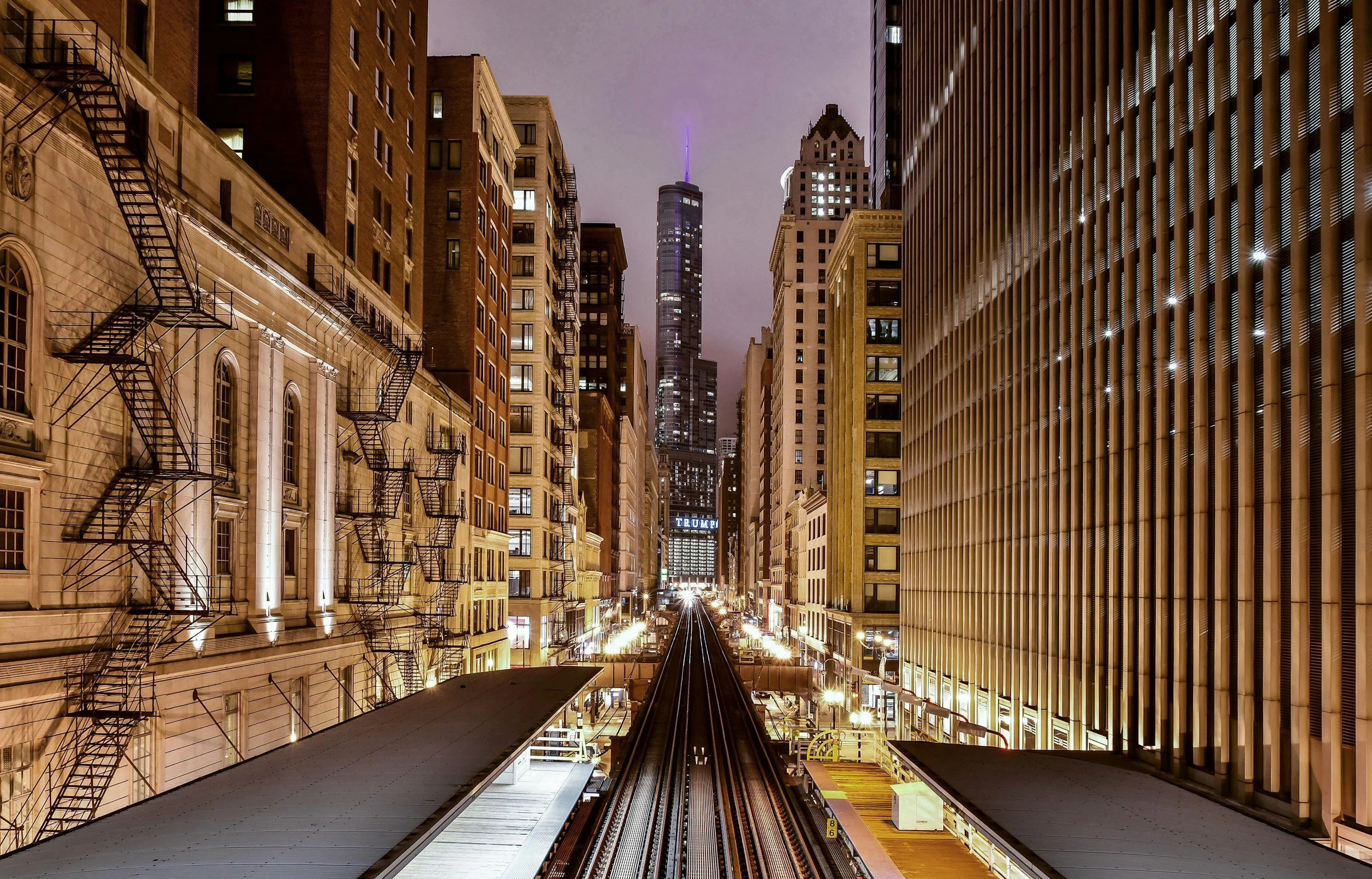 Nighttime city scene viewed from the L station platform, with buildings on either side, fire escape staircases on the left, and a tall skyscraper in the distance illuminated with purple light.